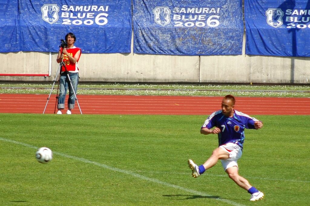 2006年の日本代表ホームシャツを着てシュートを放つ選手。背景の看板に「SAMURAI BLUE 2006」